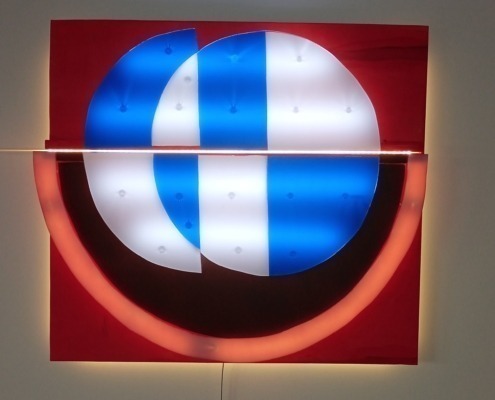 Entdecken Sie eine moderne Lichtplastik, die das Logo abstrakt interpretiert. Das Kunstwerk besteht aus einem Holz-Unterbau und mehreren Acrylglas-Scheiben, die mit weißen LEDs hinterleuchtet werden. Ideal für zeitgenössische Innenräume, Galerien und lichtbasierte Kunstinstallationen.